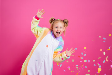 Happy little girl in kigurumi unicorn on a pink background rejoices in multi-colored confetti, space for text