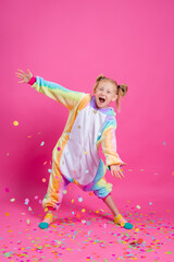 Happy little girl in kigurumi unicorn on a pink background rejoices in multi-colored confetti, space for text