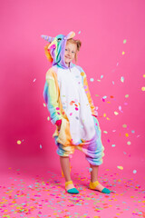 Happy little girl in kigurumi unicorn on a pink background rejoices in multi-colored confetti, space for text