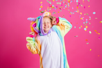 Happy little girl in kigurumi unicorn on a pink background rejoices in multi-colored confetti, space for text