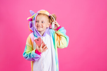 Happy little blonde girl in unicorn kigurumi listens to music holding in hand smatrphone on a pink background