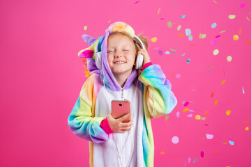 Happy little blonde girl in unicorn kigurumi listens to music holding in hand smatrphone on a pink background