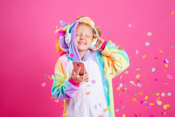 Happy little blonde girl in unicorn kigurumi listens to music holding in hand smatrphone on a pink background