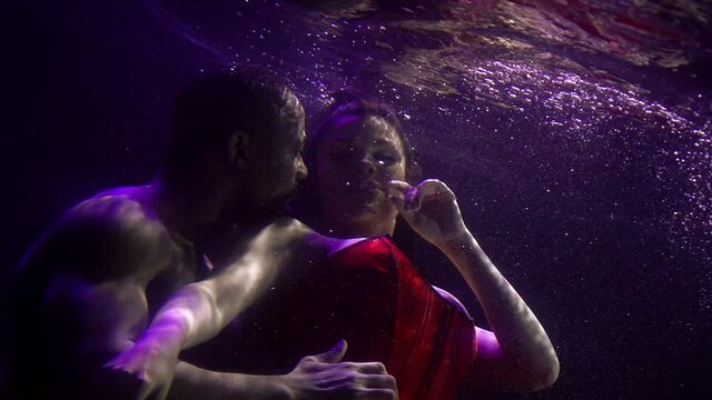 Close-up portrait of a tender couple in love who are under water and hugging, a buxom girl in a dress and a dark-skinned man. They are on a dark background with bubbles.