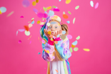Obraz na płótnie Canvas Happy little girl in kigurumi unicorn on a pink background rejoices in multi-colored confetti, space for text