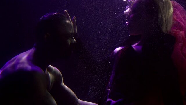 Close-up portrait of a tender couple in love who are under water and look at each other, a buxom girl in a dress and a dark-skinned man. They are on a dark background with bubbles.