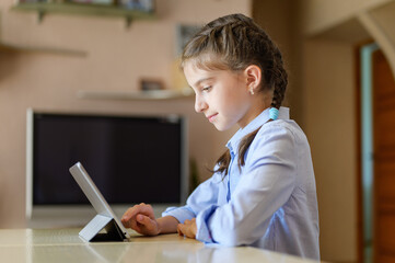 The girl at home sits at a table and uses a tablet.