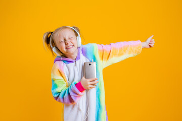 A happy little girl in a kigurumi unicorn listens to music in headphones holding a smartphone. yellow background, space for text