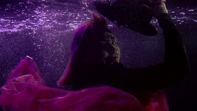 Close-up portrait of a buxom girl in a black hat and red lipstick on her lips, she is under water. It floats on a dark background with bubbles and fabric from the skirt developing around it.