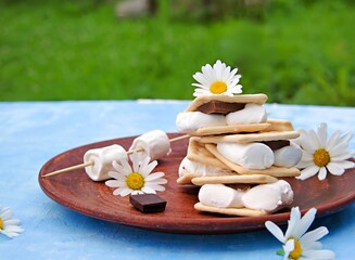 Obraz na płótnie Canvas Homemade smores dessert with marshmallows, milk chocolate and cracker on a brown clay plate on a blue concrete background.