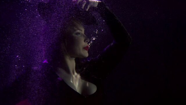 Close-up portrait of a buxom girl in a black hat and red lipstick on her lips, she is under water. It floats on a dark background with bubbles and fabric from the skirt developing around it.