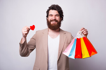 Happy cheerful man is holding a little red paper heart and some shopping bags over white background