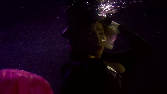 Close-up portrait of a buxom girl in a black hat, who is blowing bubbles from her mouth, she is under water. It floats on a dark background with the fabric from the skirt fluttering around it.