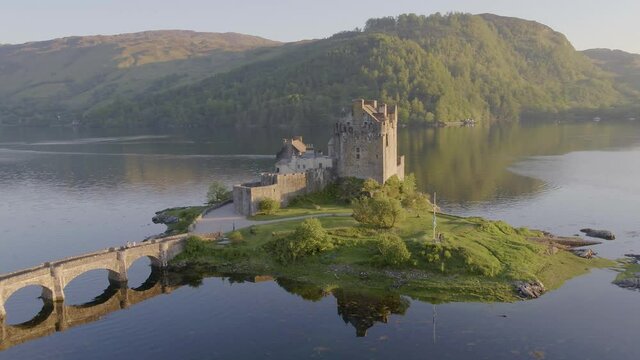 Aerial drone shot tracking around the stunning Eilean Donan Castle in Scotland, with the lake surrounding it and mountains in the background, and the reflection in the water.