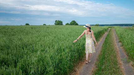 Portrait of a young woman wearing in a yellow dress and hat walking on a country road along a green wheat field.  