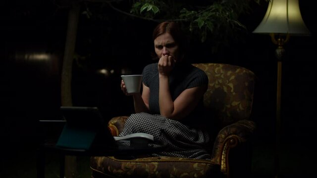 Young 30s Woman Watches Scary Movie On Tablet Computer Late At Night Sitting In Cozy Armchair With Floor Lamp In The Backyard. Woman Empathizes With Film Heroes On Gadget Screen.