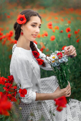 Young pretty girl Slavic or Ukrainian posing in folk dress on a flowering poppy field. Female holding a bouquet of wildflowers.