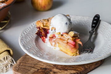 Piece of traditional apple pie with vanilla ice cream, and whole pie in ceramic baking form on white kitchen.