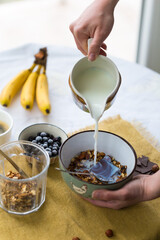 Making breakfast. Pouring milk into bowl with homemade granola with nuts, dark chocolate and bilberries.