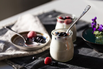 Two glass jars with natural homemade yogurt with frozen strawberries, bilberries and jam on black fabric background .