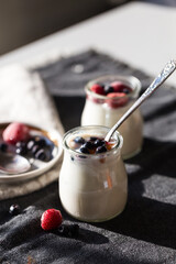 Two glass jars with natural homemade yogurt with frozen strawberries, bilberries and jam on black fabric background .