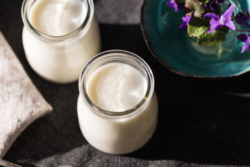 Two glass jars with natural homemade yogurt, spoons, little vase with violets on black fabric on white wooden table.