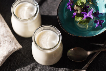 Two glass jars with natural homemade yogurt, spoons, little vase with violets on black fabric on white wooden table.