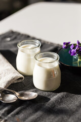 Two glass jars with natural homemade yogurt, spoons, little vase with violets on black fabric on...