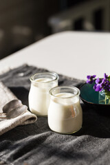 Two glass jars with natural homemade yogurt and  little vase with violets on black fabric on white wooden table.