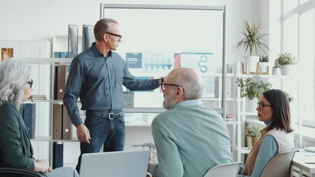 Middle-aged businessman explaining information on posters attached to glass office wall and answering questions of mixed-aged colleagues while giving presentation in the office