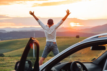 Young man having a long auto trip break. He stopped new car and funny jumping near his vehicle and enjoying the sunset orange-pink sky colors. Traveling by car and leader success concept image.