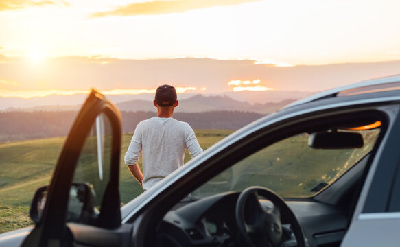 Young man having the long auto trip break. He stoped new car, standing near his vehicle and enjoying the sunset orange-pink sky colors.  Traveling by car by the empty nature vastness concept image.