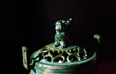 In front of a dark background is an old metal container with a lion on it, from which smoke from an incense stick rises