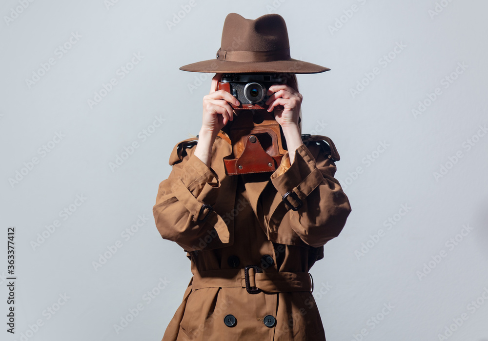 Poster woman in hat and cloak as a spy with camera on gray background.