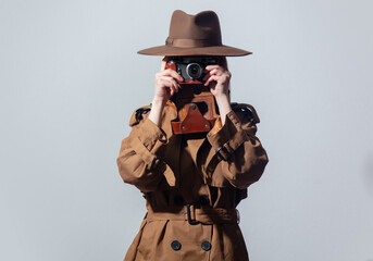 Woman in hat and cloak as a spy with camera on gray background.