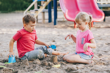 Two Caucasian children sitting in sandbox playing with beach toys. Little girl and boy friends having fun together on playground. Summer outdoors activity for kids. Leisure time lifestyle childhood.