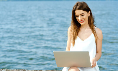 Smiling girl with laptop on her lap sits on a dock not the shore of the ocean lake