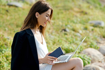 Fototapeta premium Businesswoman on a stone on the beach works behind a laptop in a white dress and black jacket 