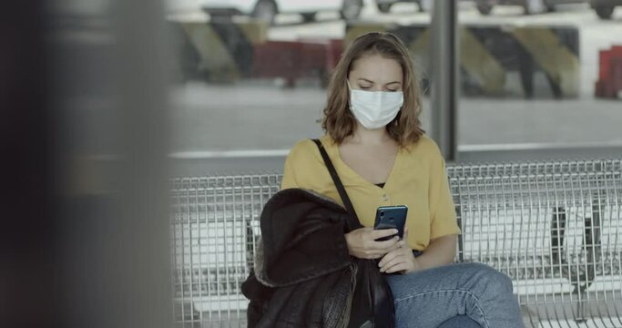 Pretty female in airport bus terminal wearing protective face mask using smart phone during covid19 pandemic