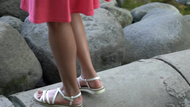 Close up of an attractive mixed race African American woman's feet in casual white strapped sandals and pink skirt as she climbs on top of a concrete storm sewer pipe and gravel path along a river.