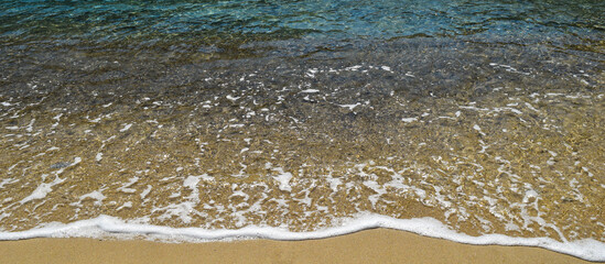 Fototapeta na wymiar Soft waves with foam on crystal clear transparent shoreline on the shore of a paradise beach in Ikaria, Aegean Sea in Greece