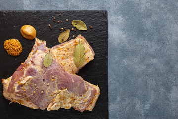 Two slices of raw lard with a layer of meat, with mustard, pepper and bay leaf, against a dark background