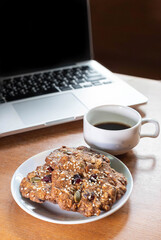 Cookie made by Grain , Raisin ,Almond, Pumpkin Seed, Cashew Nut, Cranberry , Walnut, Sunflower Seed, Chia Seed,Black Sesame Seed with a background of laptop and a cup of black coffee on wooden table