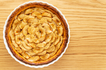 Homemade baked French apple tart, an open faced apple pie, in a baking white ceramic dish on an oak wood background. Flat lay, top view with copy space.
