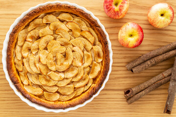 Homemade baked French apple tart, an open faced apple pie, in a baking white ceramic dish aside Gala red apples and cinnamon sticks, all on an oak wood background. Flat lay, top view.