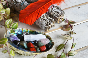An image of several tumbled healing crystals in an abalone shell with white sage bundles and sacred feather. 