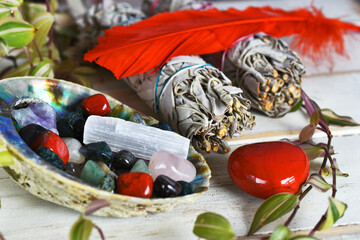 An image of several tumbled healing crystals in an abalone shell with white sage bundles and sacred feather. 