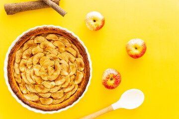 Homemade baked French apple tart, an open faced apple pie, in a baking white ceramic dish aside Gala apples, cooking spoon and cinnamon sticks on a yellow background. Flat lay, top view.