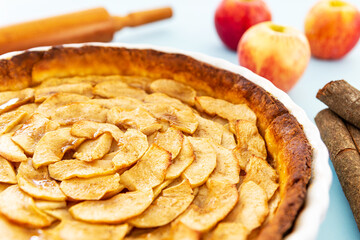 Closeup on a homemade baked French apple tart, an open faced apple pie, in a baking white ceramic dish aside Gala apples, cinnamon sticks and a rolling pin on a light blue background. Side view.