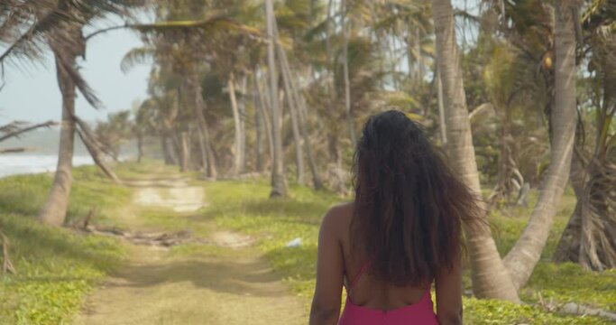 A sexy model walks along a path with coconut trees wearing a dress and her hair flowing in the wind